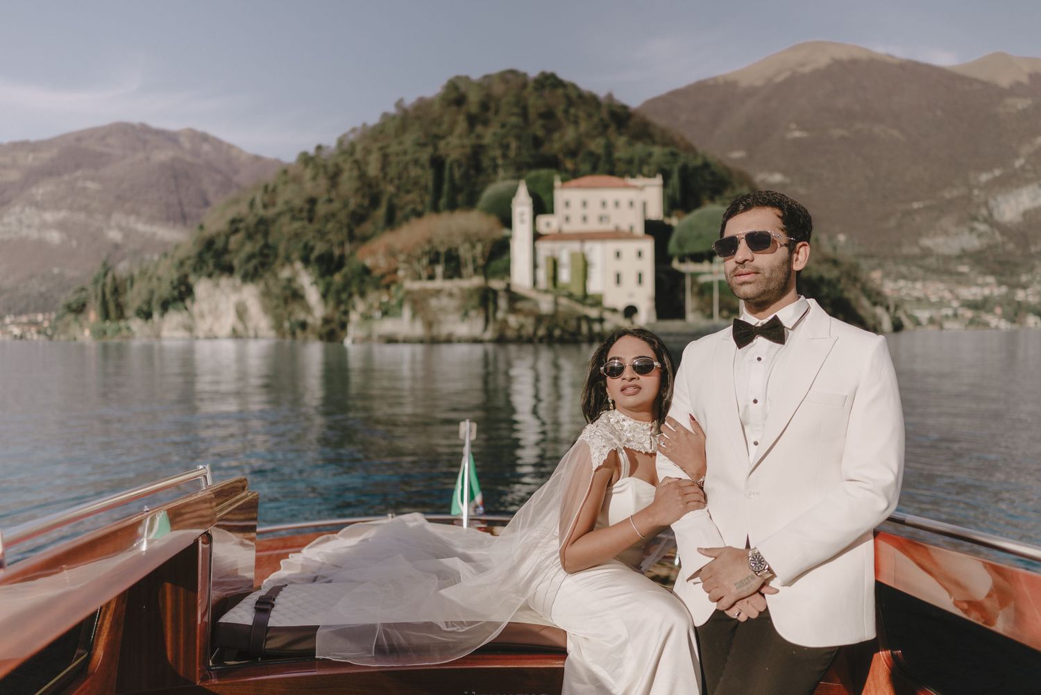 Elegant couple in wedding attire on a wooden boat on Lake Como, Italy, with a historic villa and mountains in the background.