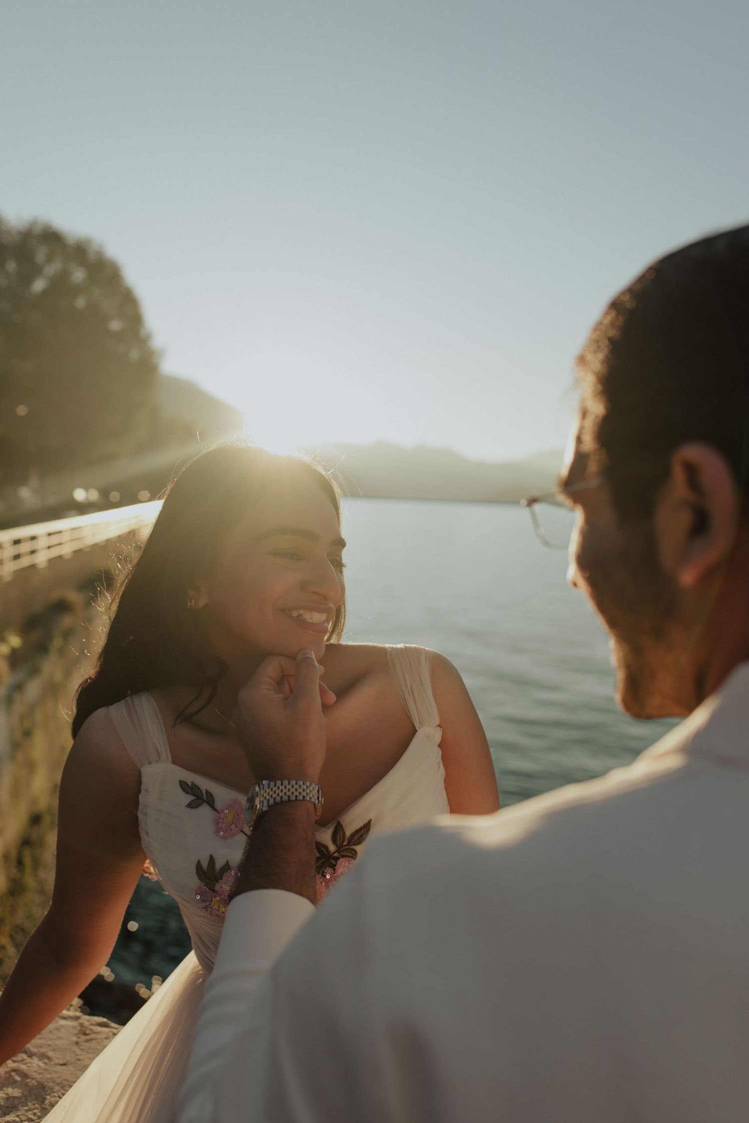 Couple sharing a romantic moment by the water at golden hour, woman smiling warmly in a white dress.