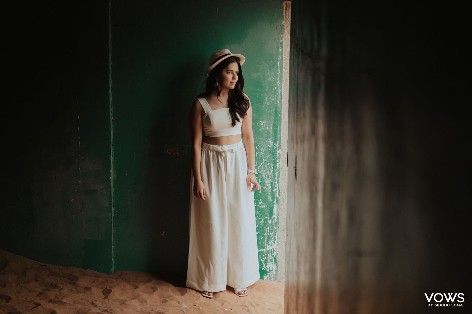 Woman in white crop top and maxi skirt with straw hat poses elegantly against a rustic green wall.