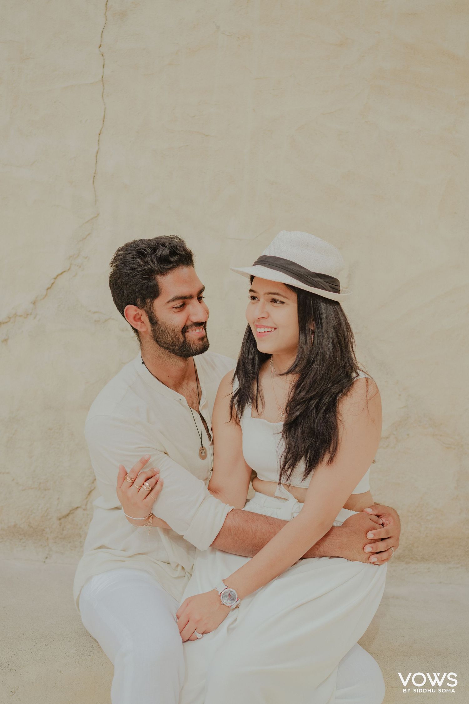 A couple in white outfits share a romantic moment, woman in panama hat, against a beige wall.