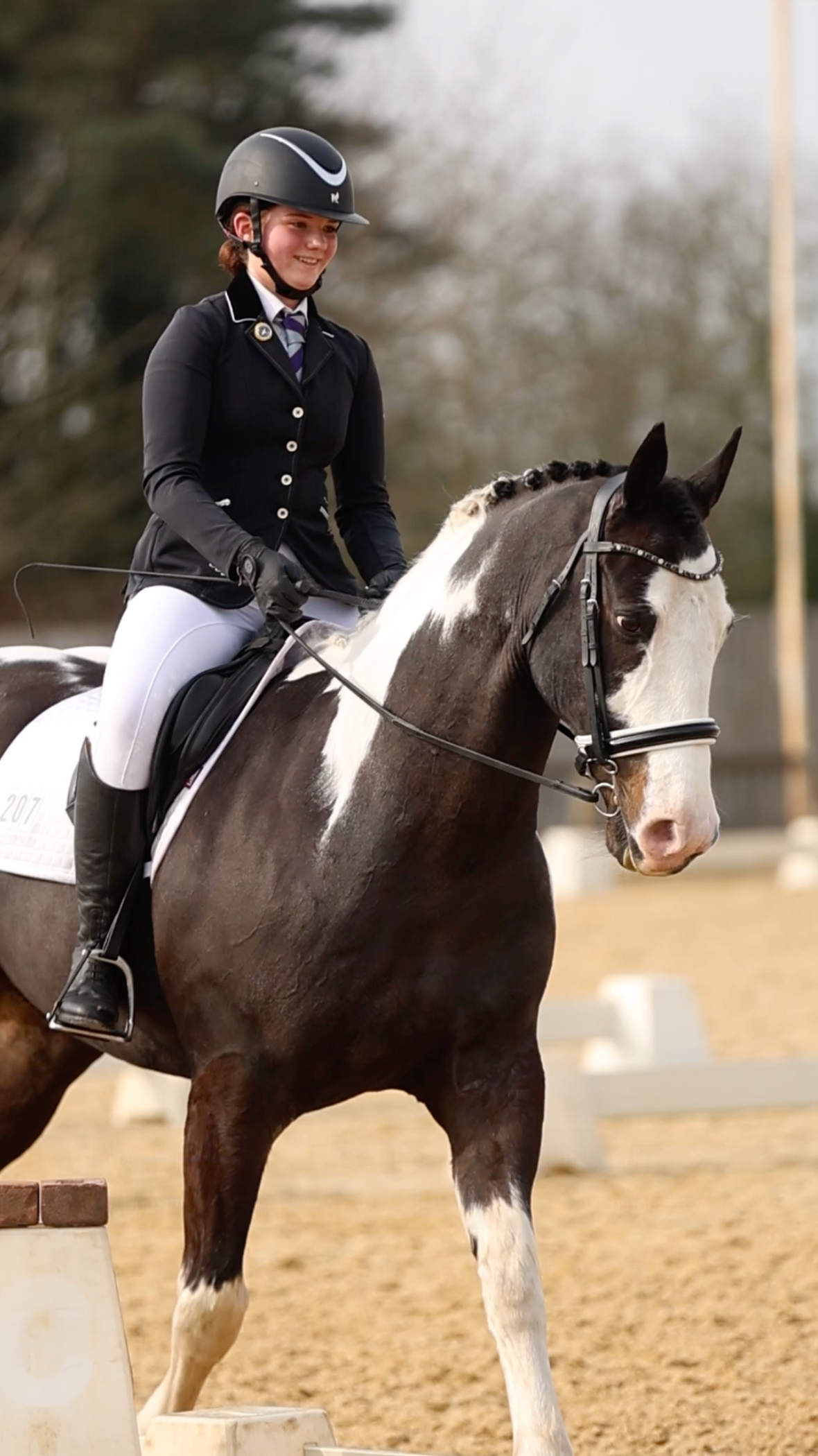 A smiling rider in black jacket and helmet performs dressage on a black and white pinto horse in an outdoor arena.