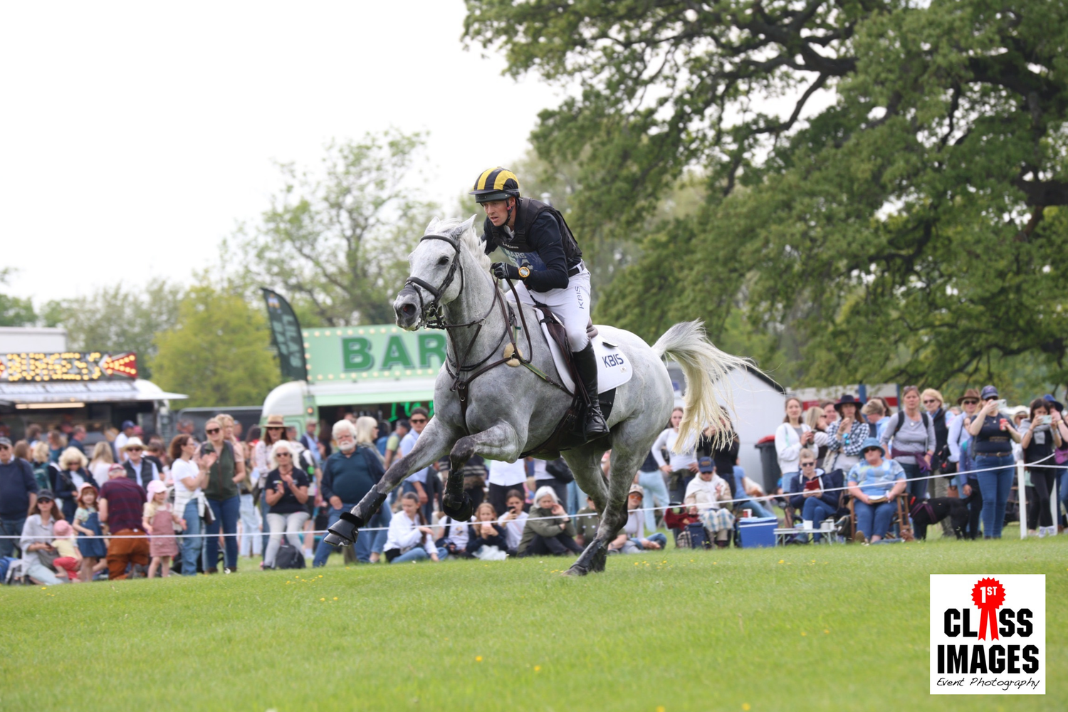 Rider on a grey horse gallops at speed during a cross-country equestrian competition event.