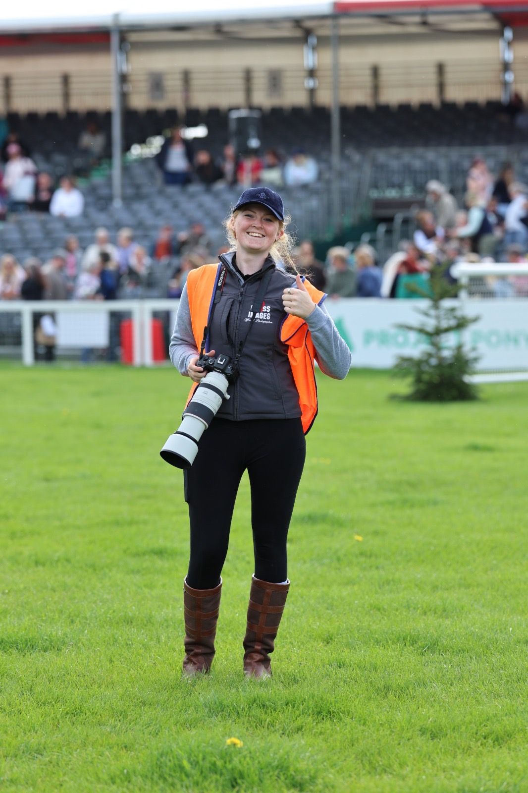 A smiling female photographer in orange safety vest gives thumbs up while holding a professional camera at an equestrian event.
