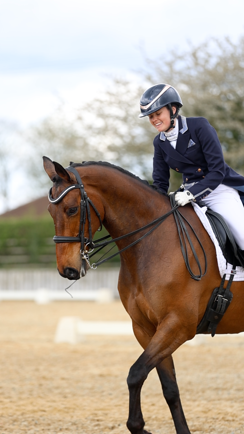 Female dressage rider in navy jacket smiling while riding bay horse in outdoor arena.