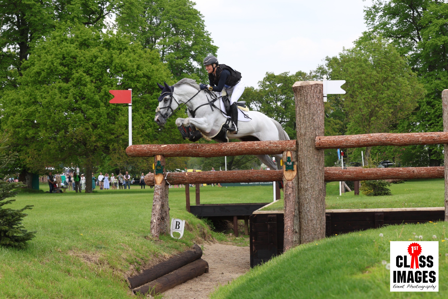 Equestrian rider on a grey horse clearing a rustic wooden cross-country fence outdoors.