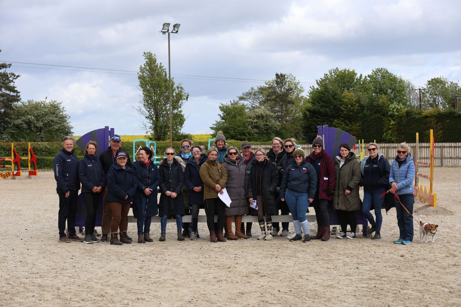 Large group of equestrian enthusiasts posing together in a sandy outdoor riding arena.