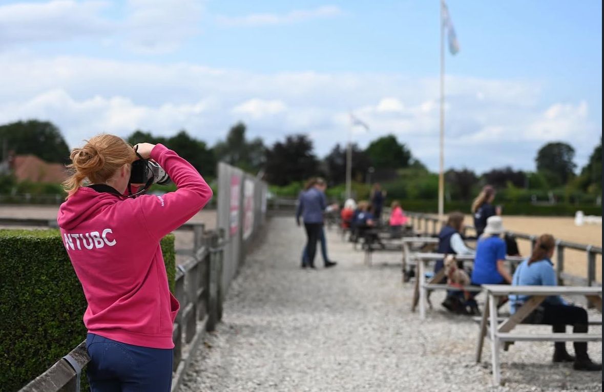 Woman in pink NTUDC hoodie watching equestrian activity at outdoor competition venue.