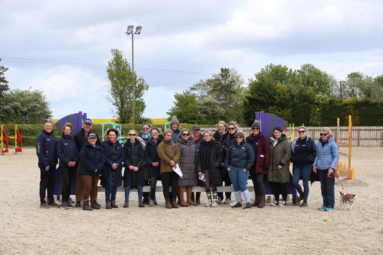 Large group of equestrian riders and coaches posing together at outdoor riding arena.