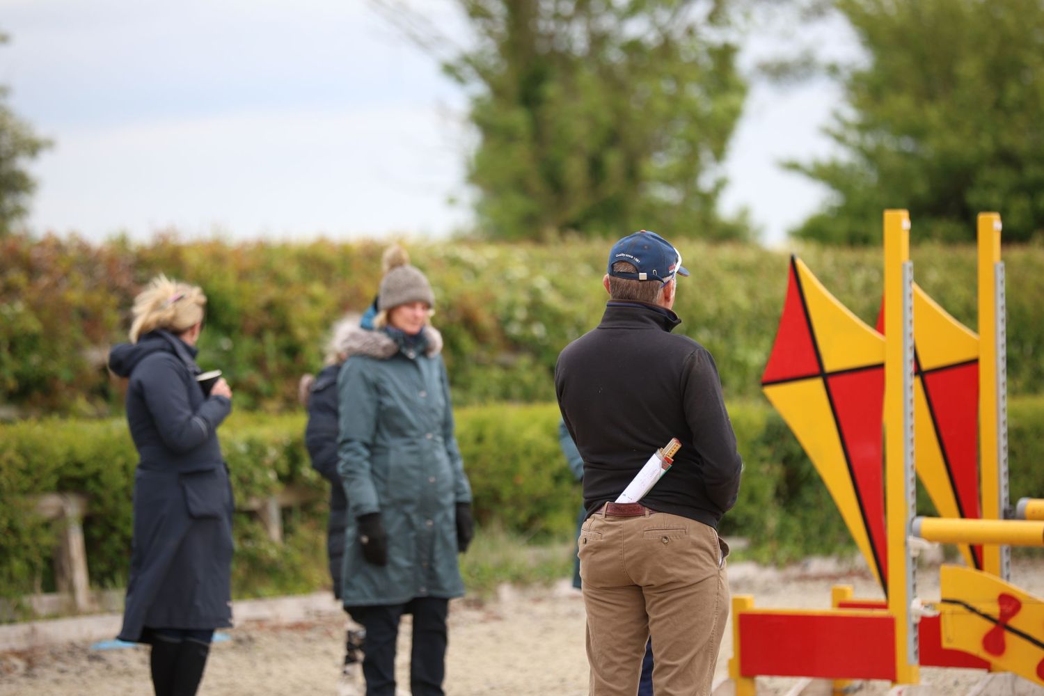 Equestrian coach observing riders near colorful show jumping fence at outdoor arena.