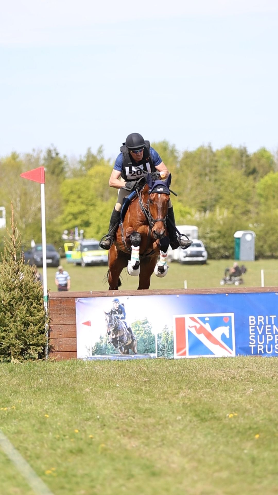Equestrian rider jumping brown horse over cross-country fence at British Eventing competition outdoors.