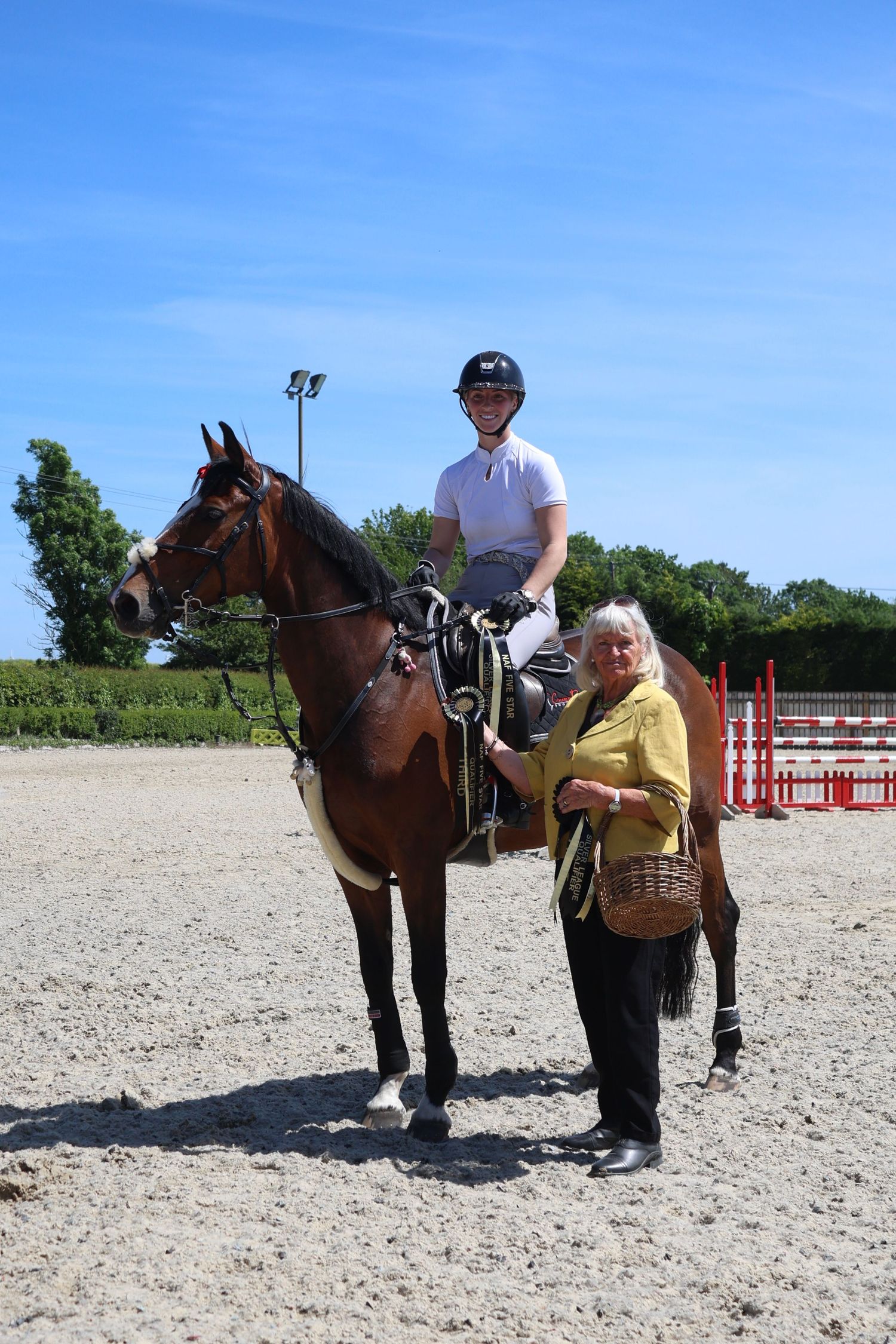 A rider in white shirt and helmet sits on a bay horse beside an older woman holding a basket at an equestrian venue.