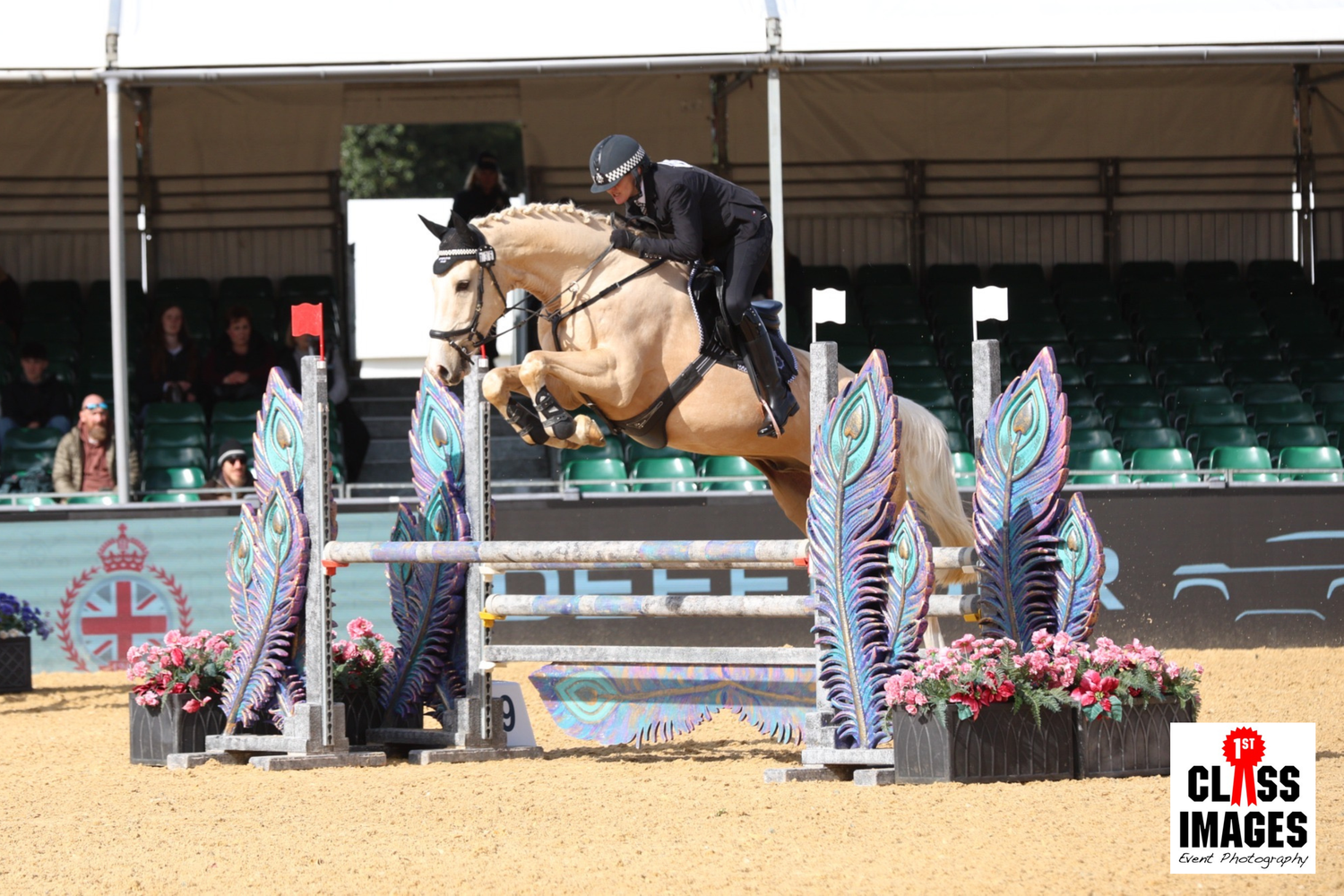 Rider on a palomino horse jumping over decorated show jumping poles in a large equestrian arena.