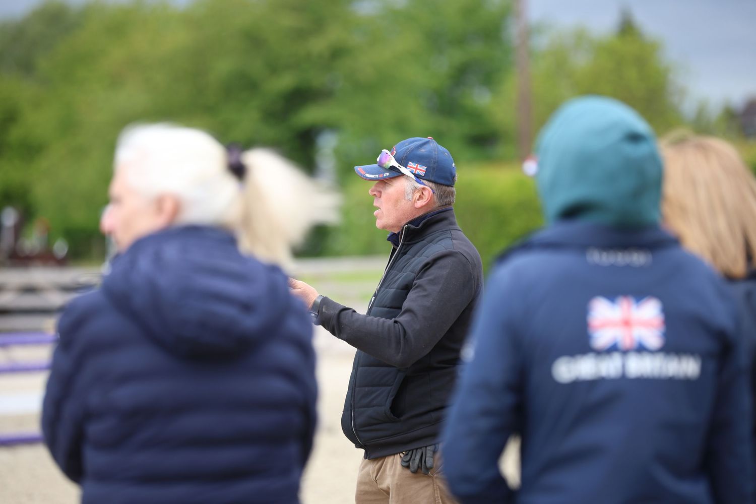 Equestrian coach in blue cap observing Team Great Britain riders at training session.