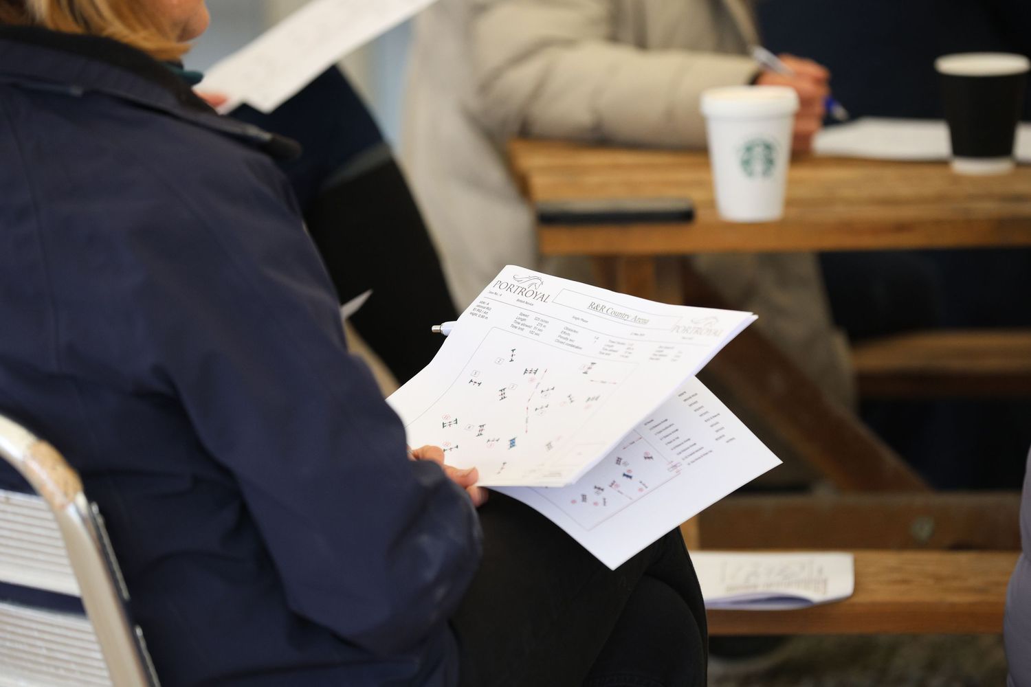 Person reviewing equestrian event paperwork at table with Starbucks coffee cup nearby.