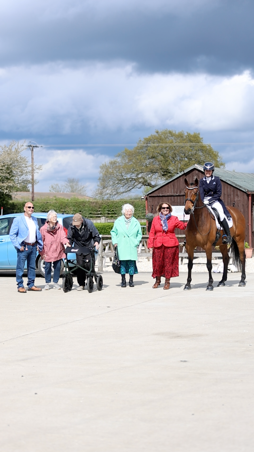 Dressage rider on bay horse greeted by group of spectators at equestrian competition yard.