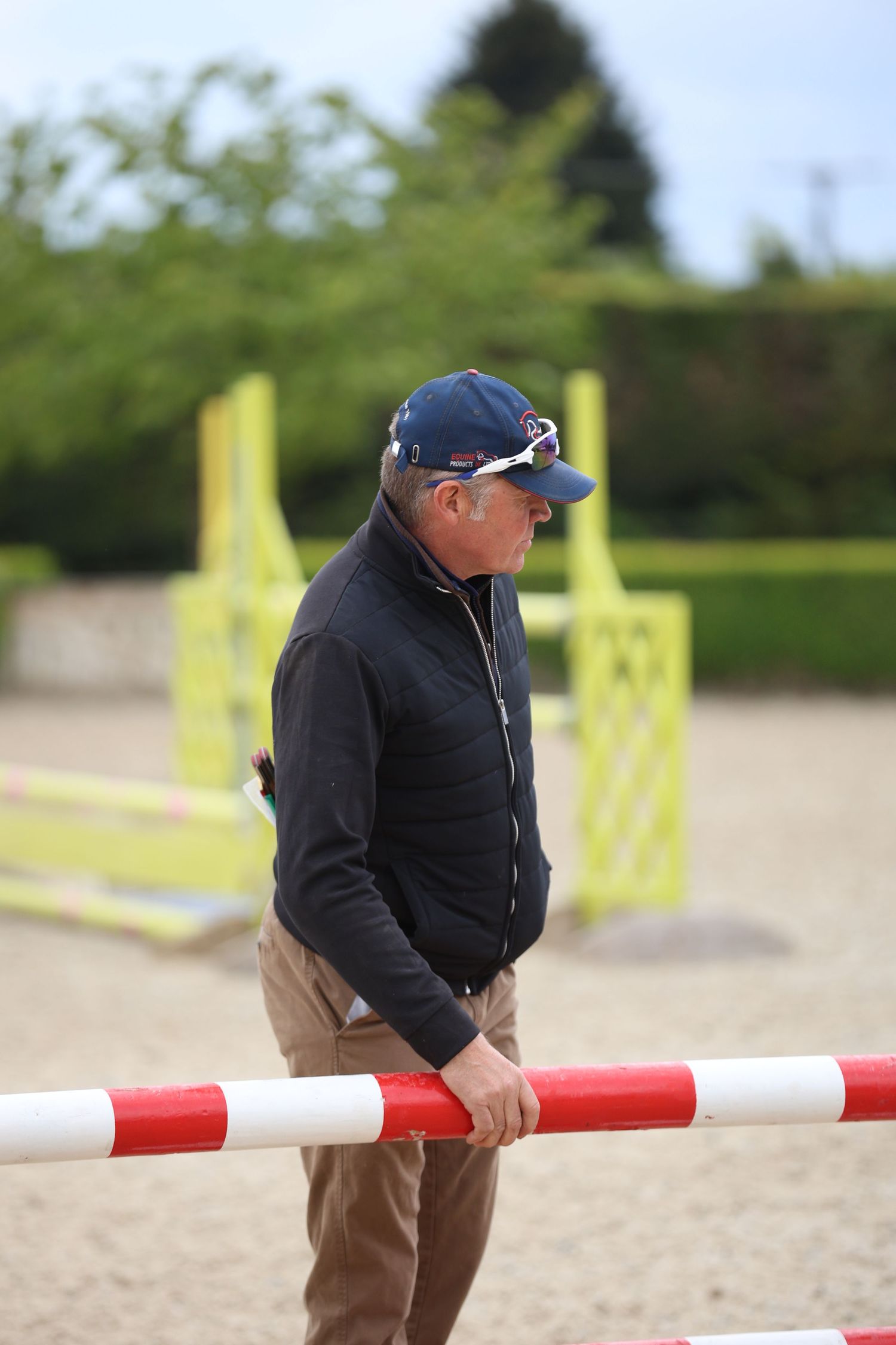 Male equestrian coach holding red and white jump pole at outdoor riding arena.