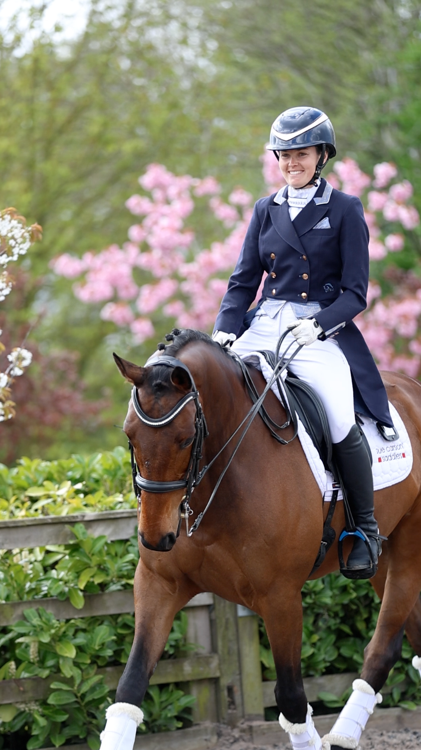 Smiling equestrian in navy dressage attire riding bay horse past pink flowering shrubs.