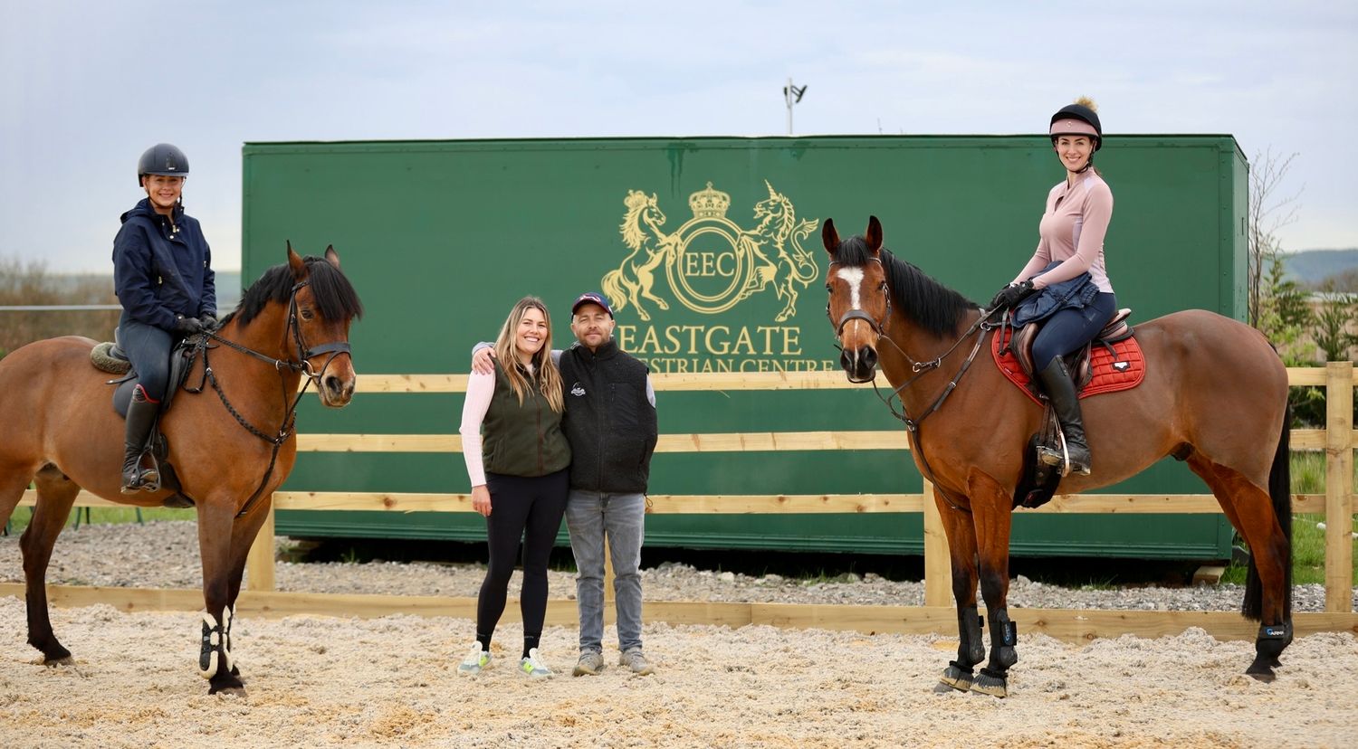 Two riders on bay horses flanking two people standing before Eastgate Equestrian Centre trailer.