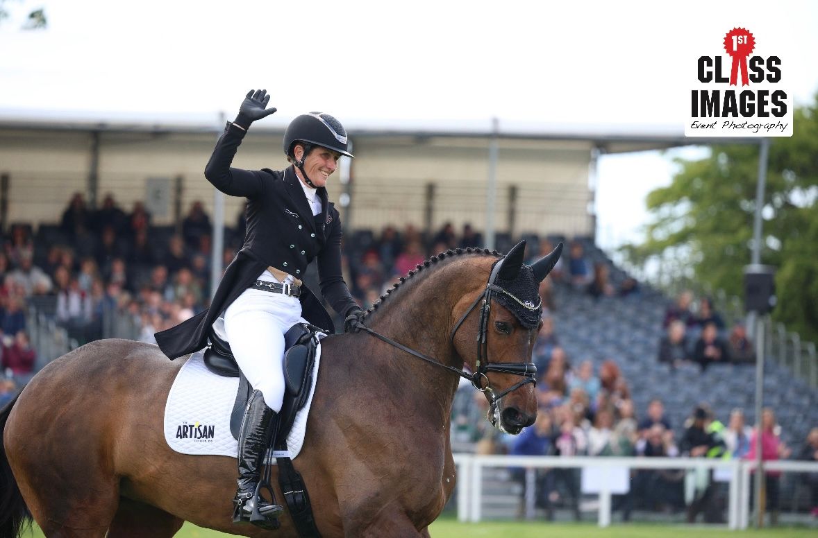 Smiling female equestrian rider waving to crowd while mounted on a chestnut horse in an arena.