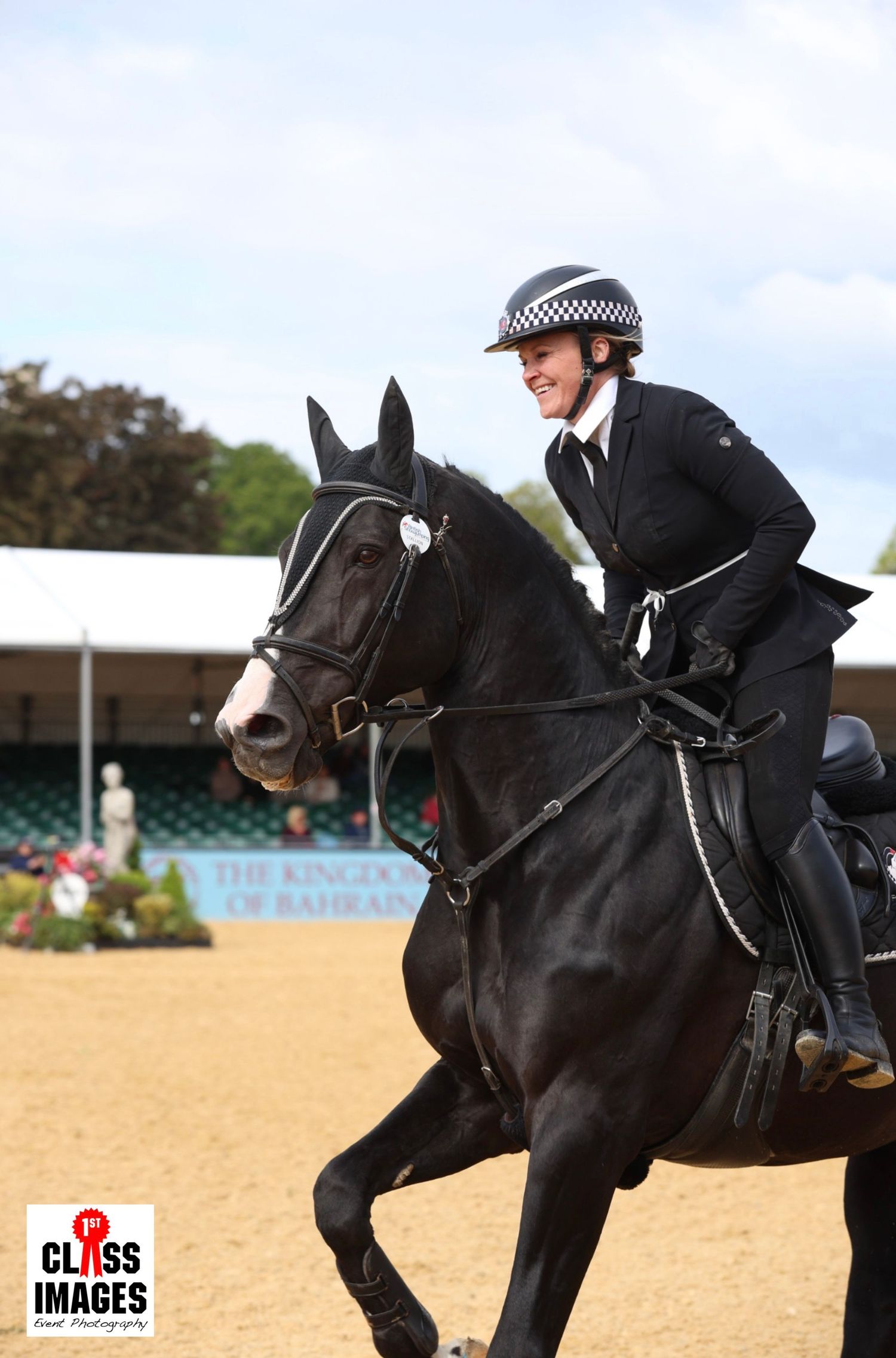 Smiling female rider in checkered helmet rides a black horse at The Kingdom of Bahrain equestrian event.