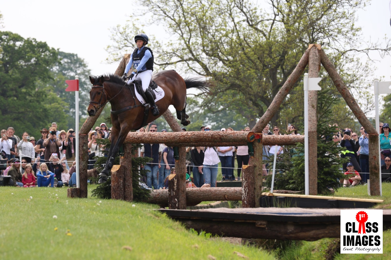 Horse and rider tackle a challenging wooden cross-country fence obstacle watched by large crowd.