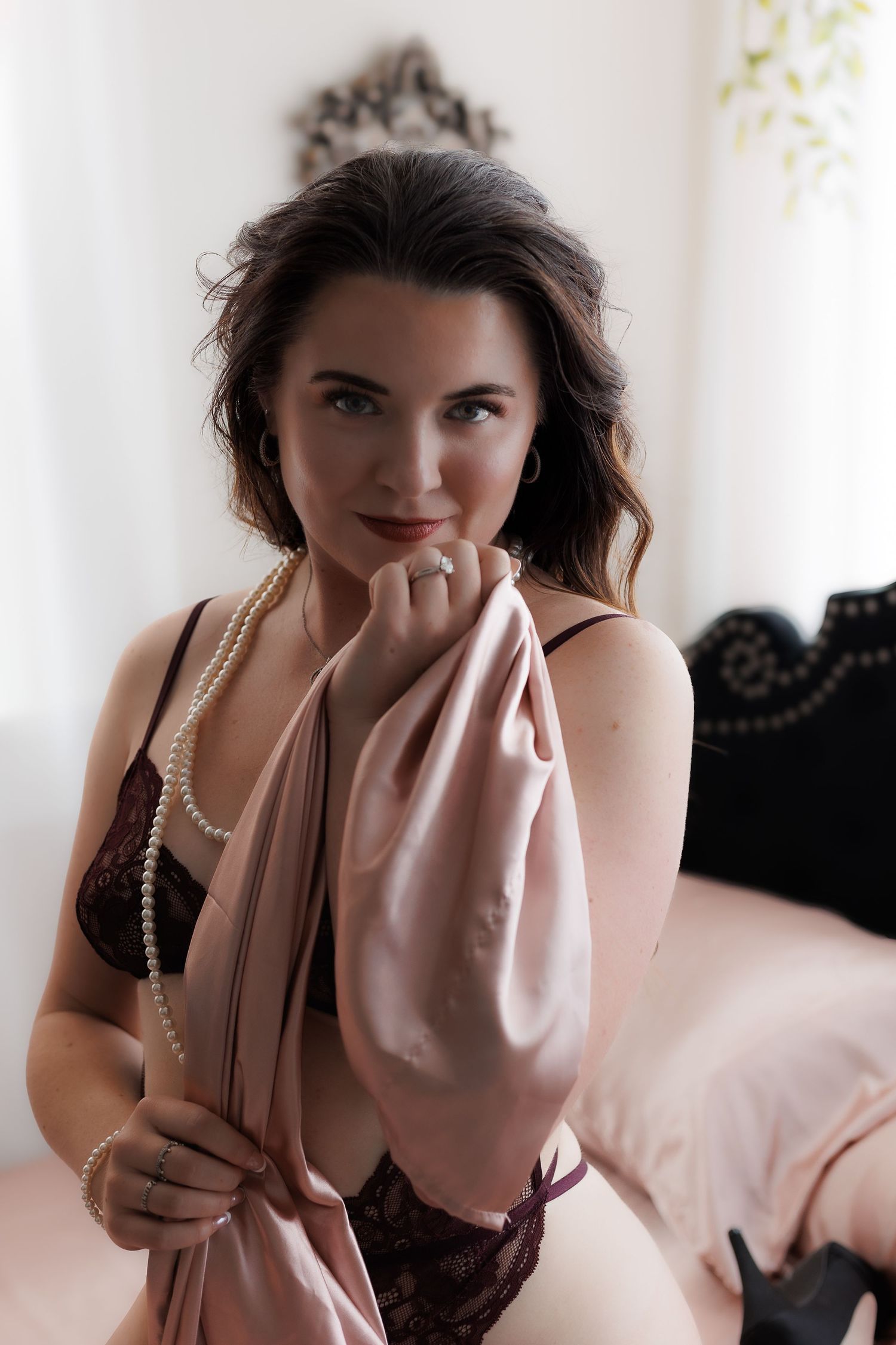 Brunette woman in lace lingerie holding pink silk robe with pearls.