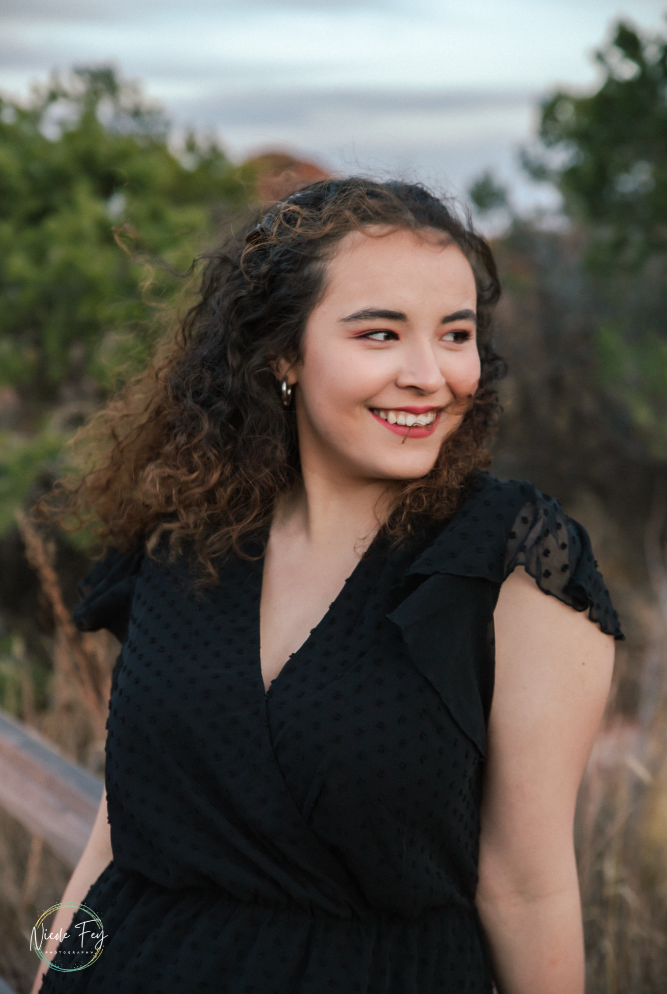 A young woman with curly hair smiles outdoors, wearing a black dress, with greenery in the background.