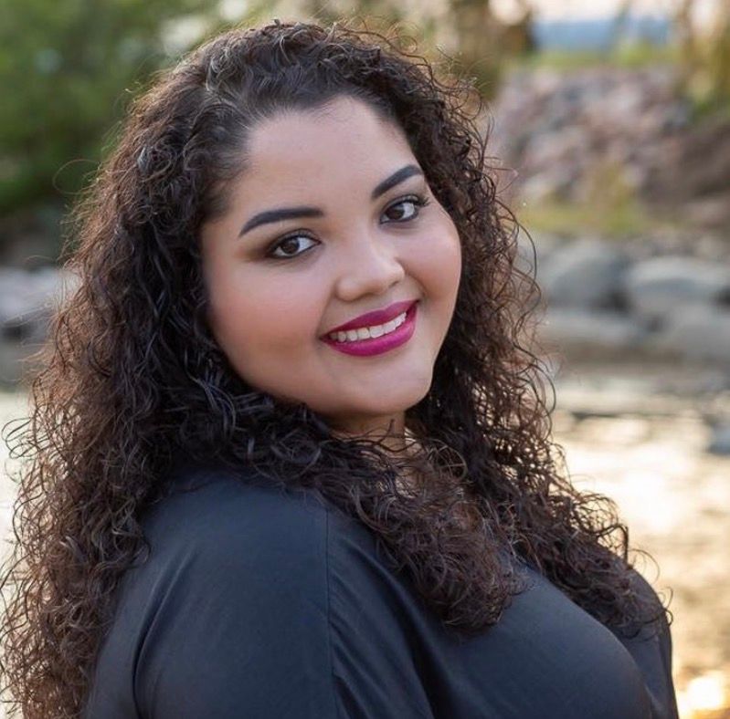 Young woman with curly dark hair and pink lips smiling outdoors in a black top with a natural background.