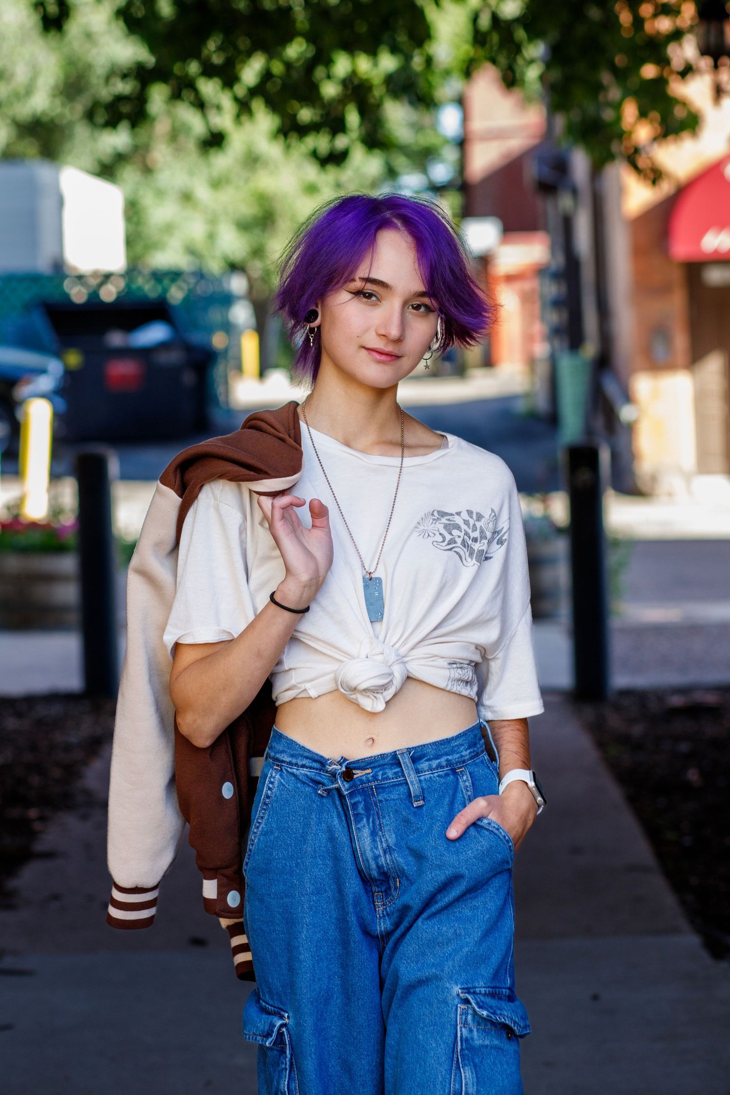 Young woman with purple hair wearing knotted white tee, jeans, and varsity jacket on urban sidewalk.