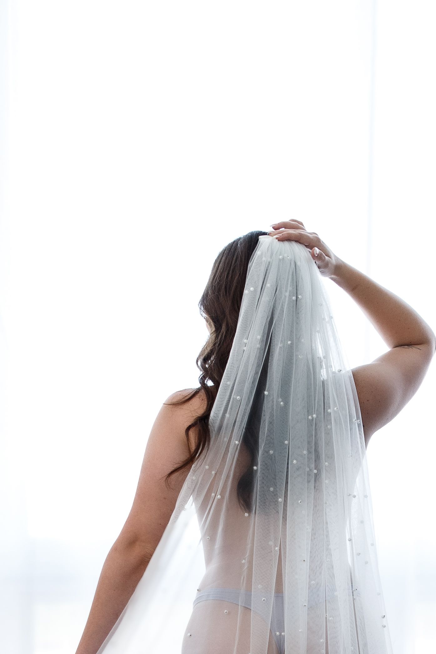 Bride seen from behind wearing a pearl-embellished wedding veil, adjusting it near a bright window.