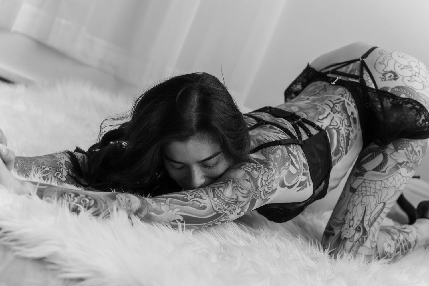 Black and white photo of a tattooed woman in lingerie lying on a fluffy white surface.