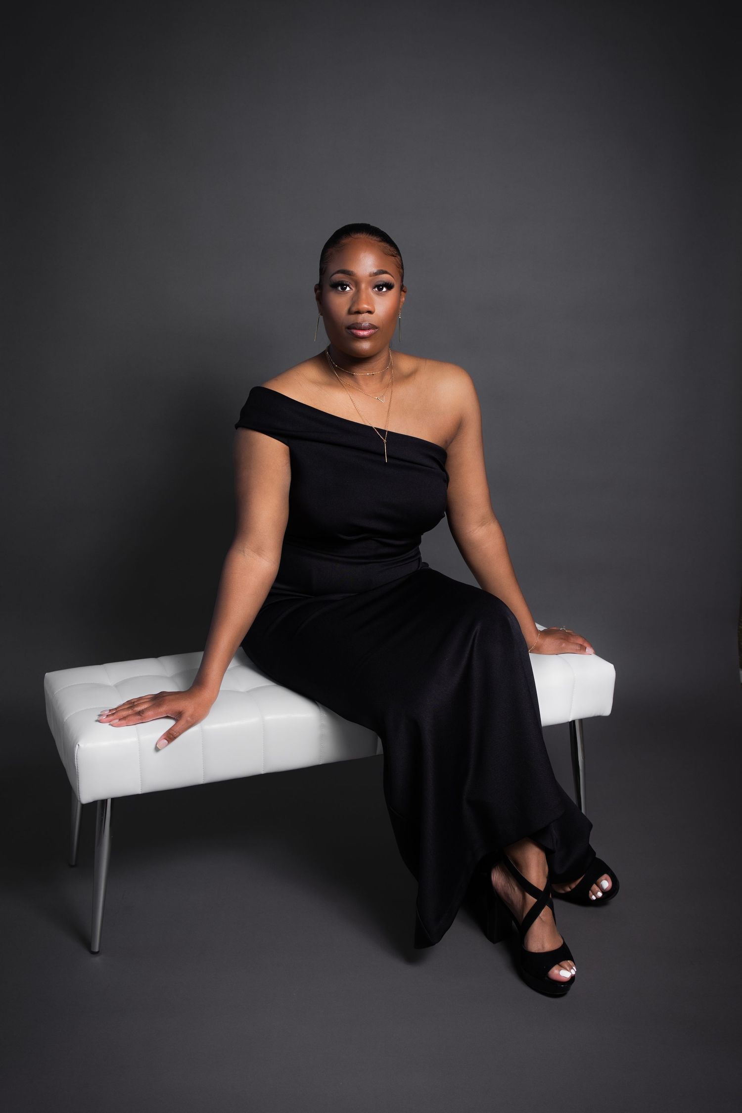 A woman in an elegant black off-shoulder gown sits gracefully on a white bench against a dark gray studio background.