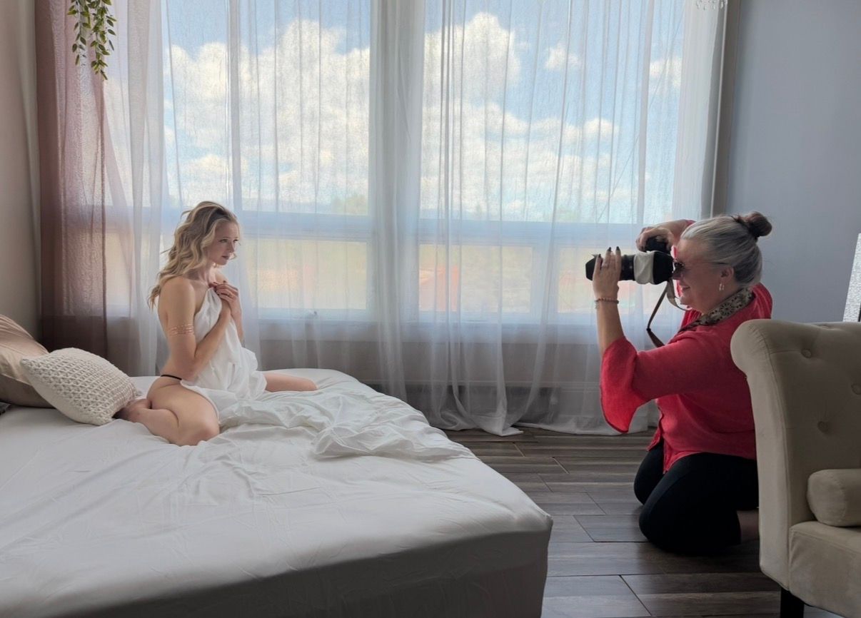Photographer in a red top crouches by a bright window to photograph a woman in white posing on a bed in a sunlit bedroom.