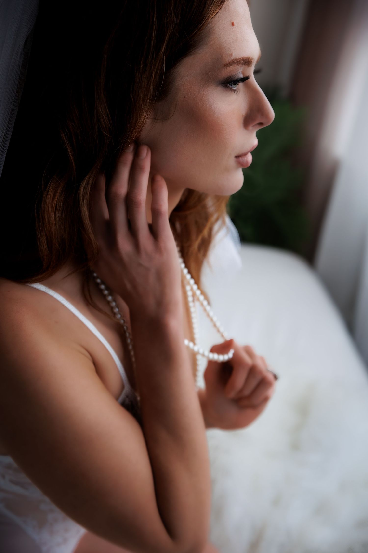 A redheaded woman in white lingerie holds pearl necklace while gazing thoughtfully to the side in soft natural light.