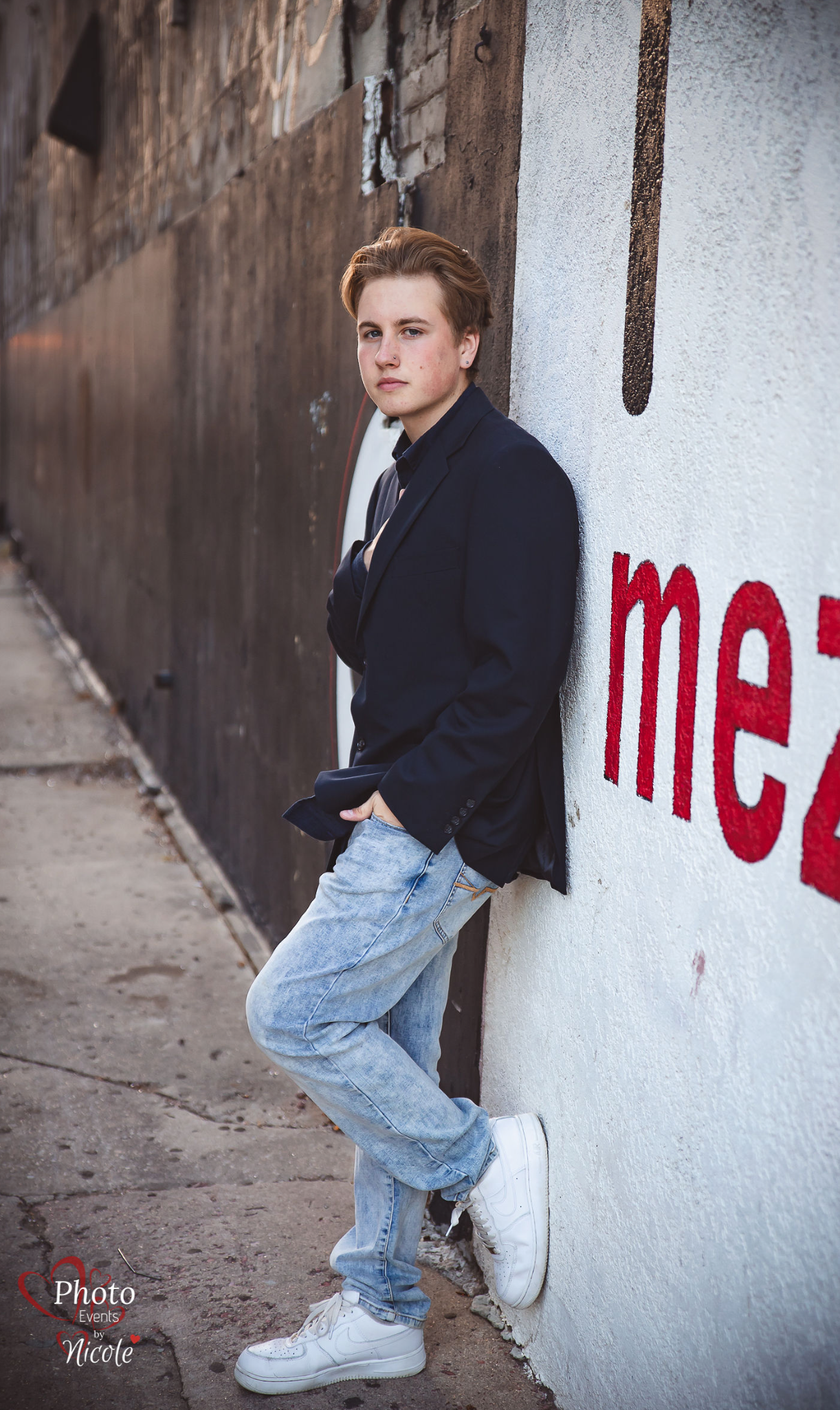 Young man in navy blazer and light jeans leans against graffiti wall in urban alley for senior portrait.