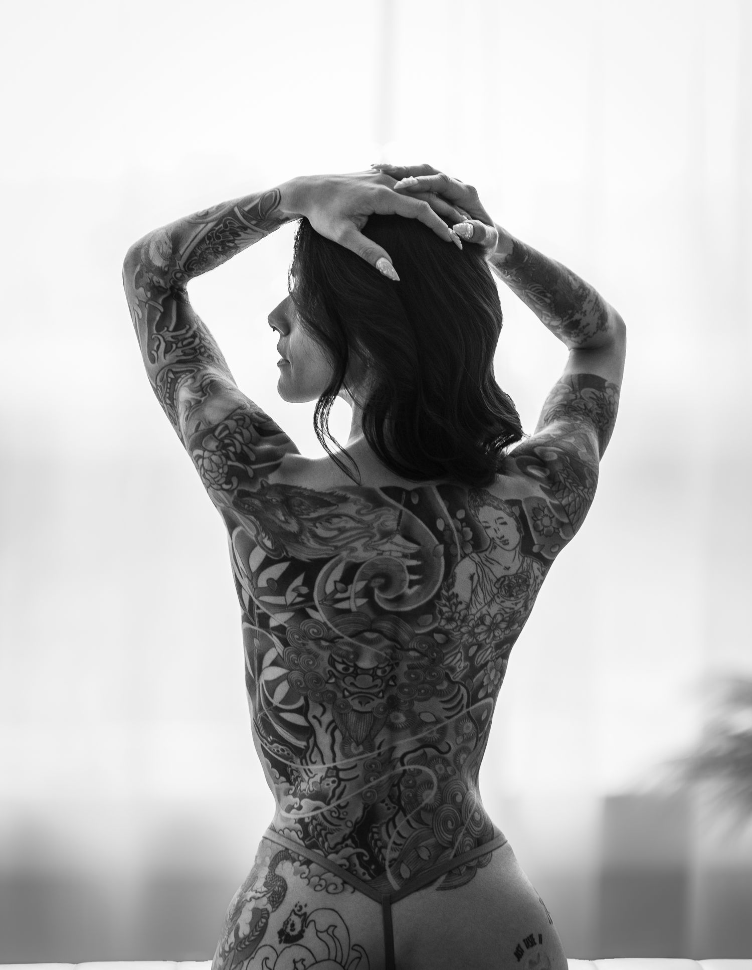 Black and white photo of a tattooed woman posing with hands on head, showcasing intricate back and sleeve tattoos.