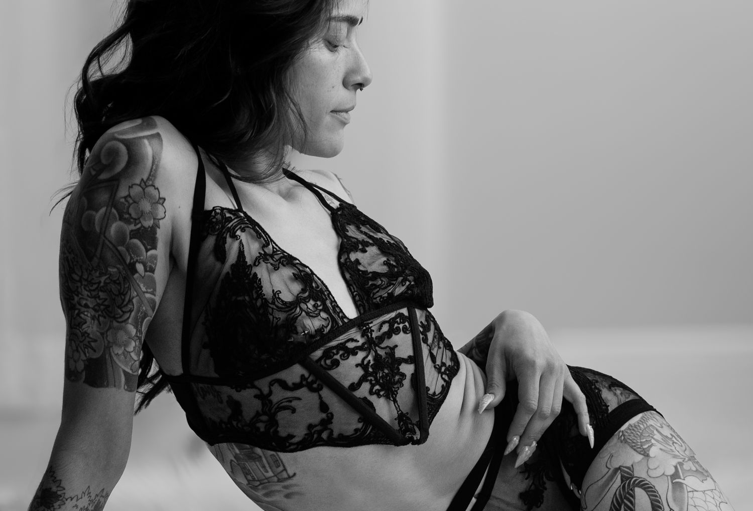 Black and white photo of a tattooed woman posing in black lace lingerie, showcasing intricate sleeve tattoos.