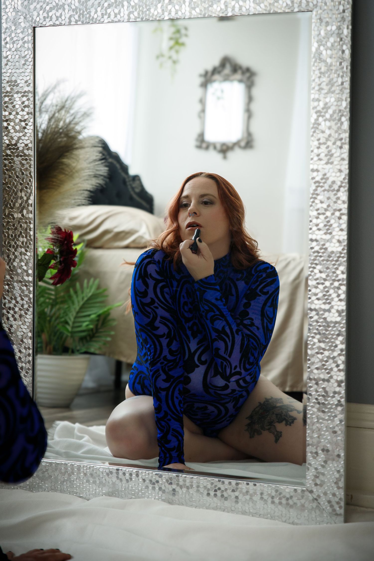 Redheaded woman in blue patterned bodysuit applying lipstick before a large ornate mirror during boudoir photography in Colorado Springs, CO