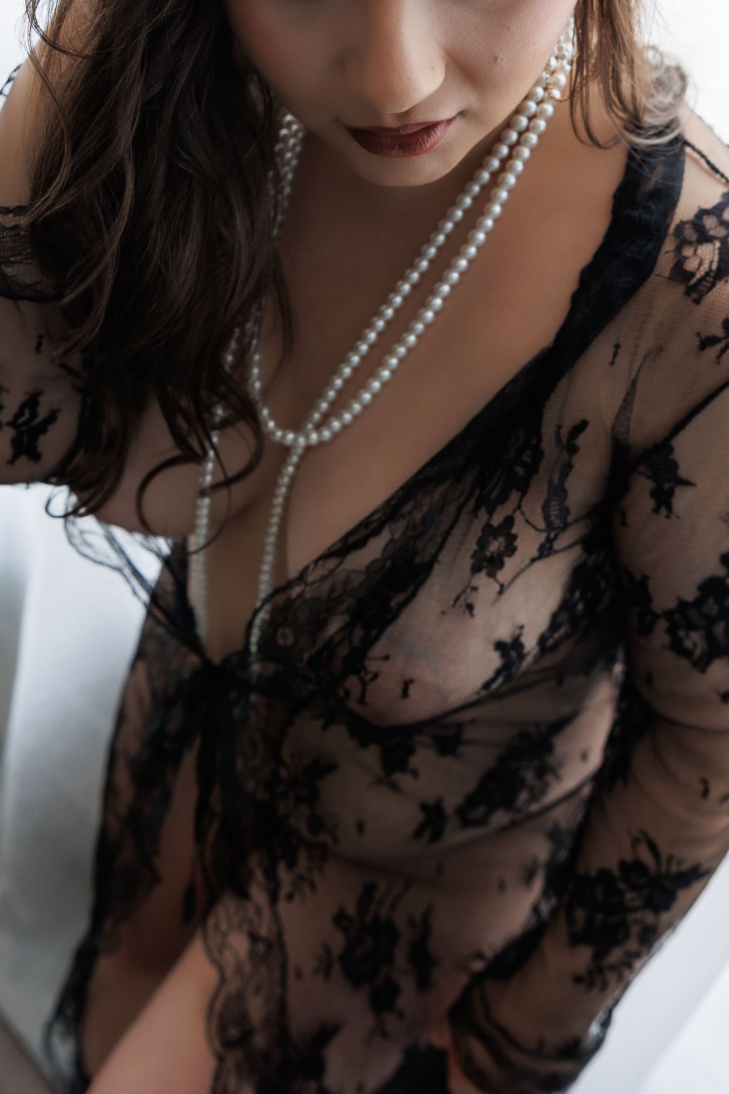 Woman in sheer black lace robe with pearl necklace posing elegantly.