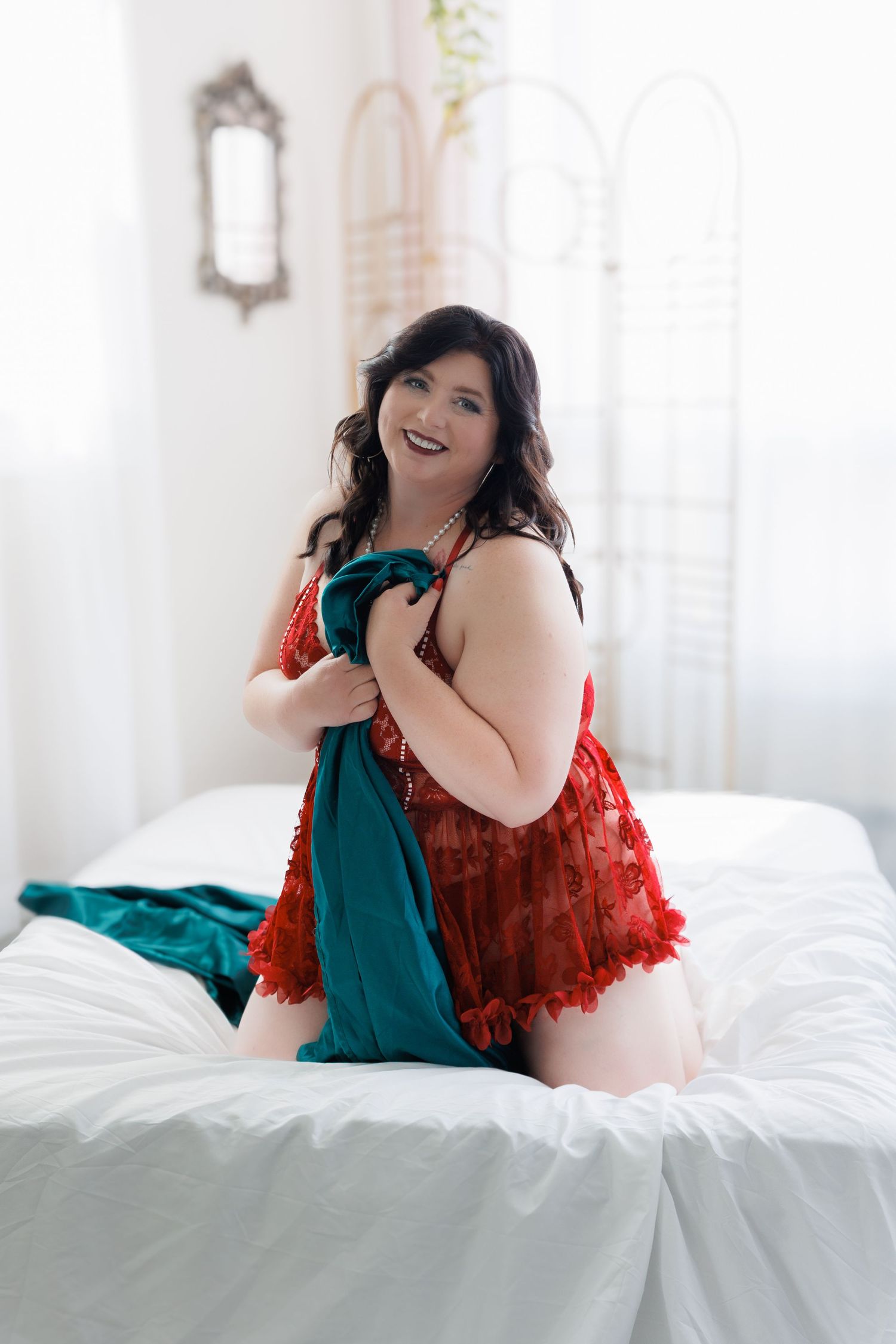 A smiling plus-size woman in red lace lingerie kneels on a white bed holding a teal fabric in a bright studio.