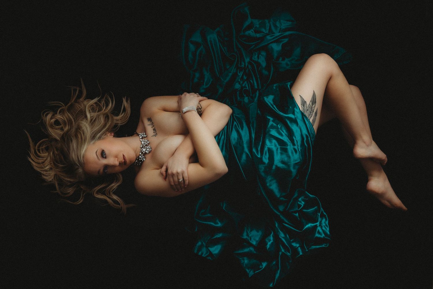 A woman with blonde hair reclines gracefully in a teal satin gown against a dark background.
