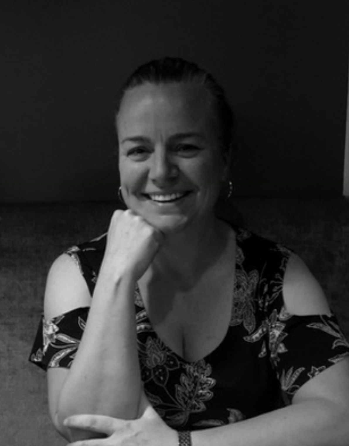 Black and white portrait of a smiling woman resting her chin on her hand, wearing a floral top.