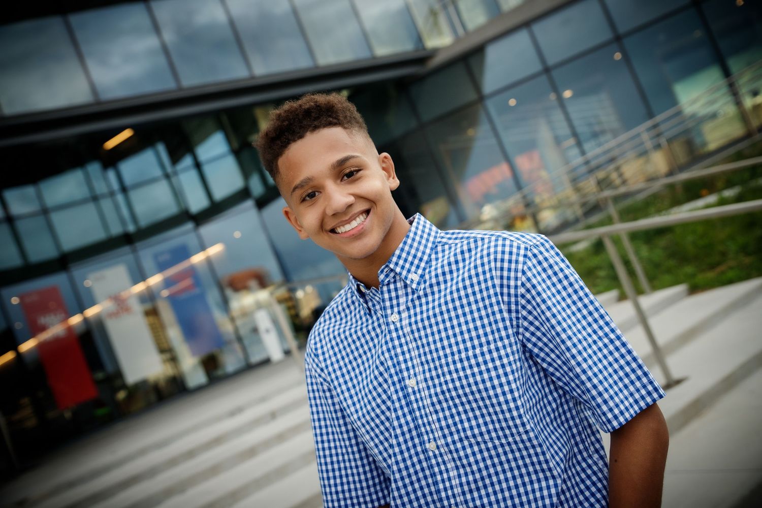 Smiling young boy in blue plaid shirt posing outdoors near modern glass building on a cloudy day.