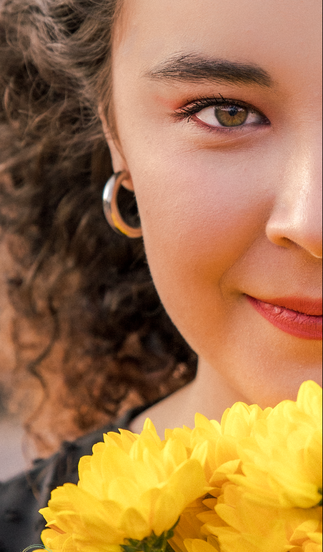 Close-up of a young woman with curly hair and hoop earrings holding bright yellow flowers, smiling softly.