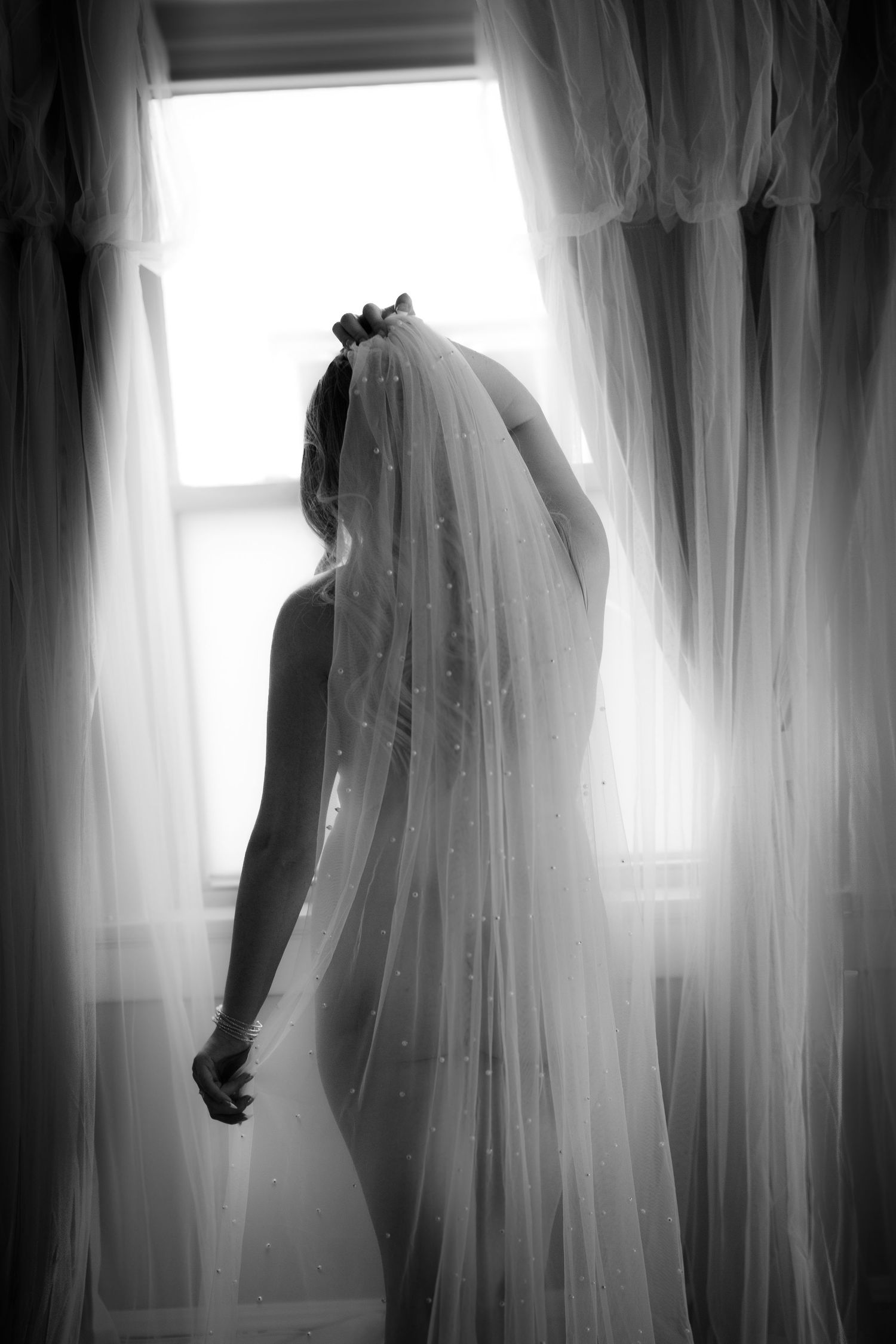 Black and white photo of a bride in a flowing veil standing by a sunlit window on her wedding day.