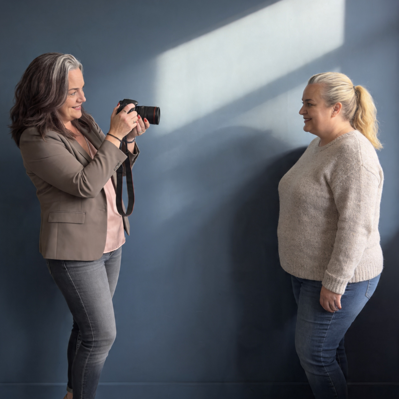 A female photographer takes a portrait photo of a smiling woman leaning against a dark blue studio wall.