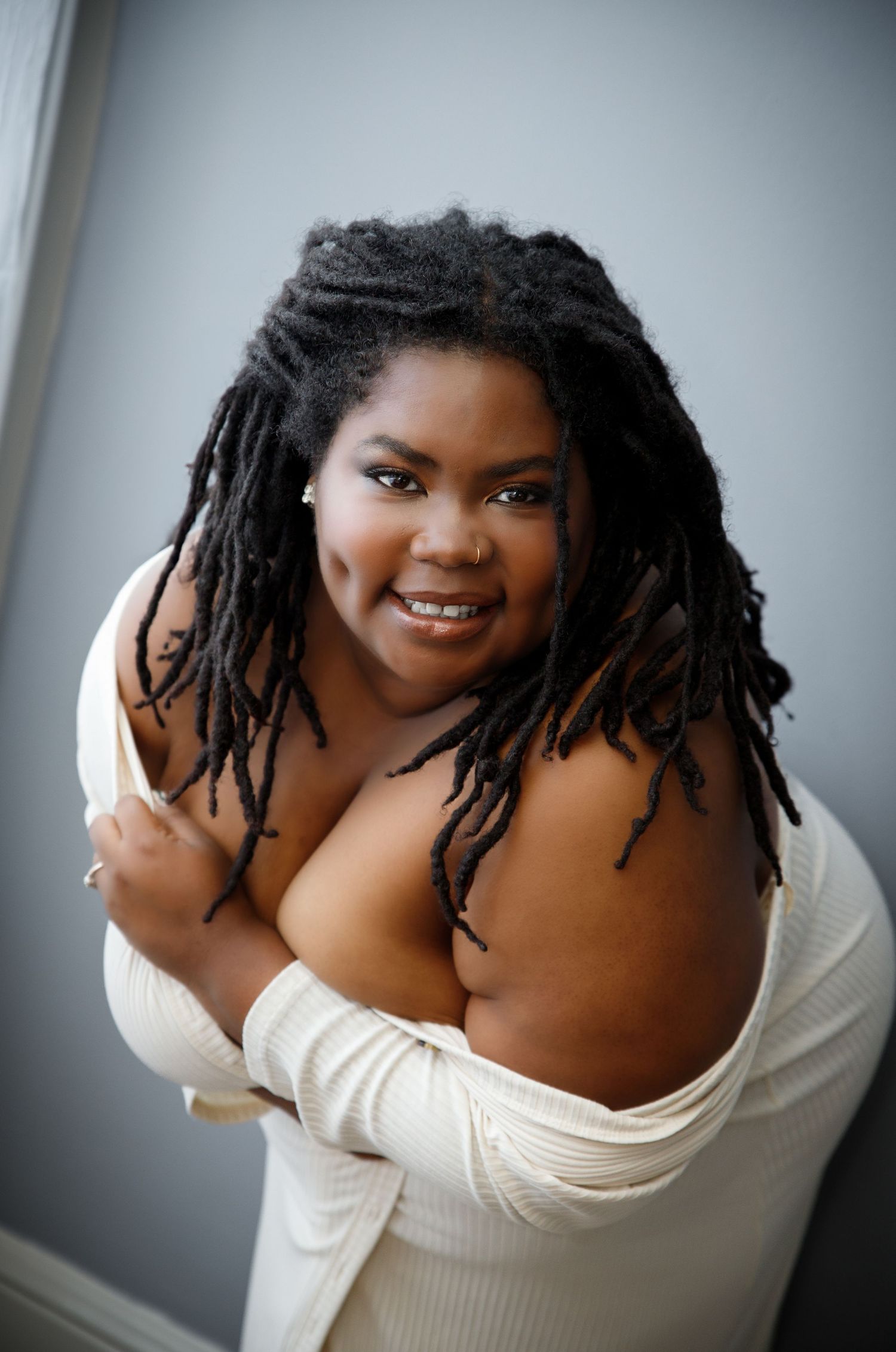 A plus-size woman with locs smiles confidently in an off-shoulder cream ribbed top at a private boudoir studio in Colorado Springs.