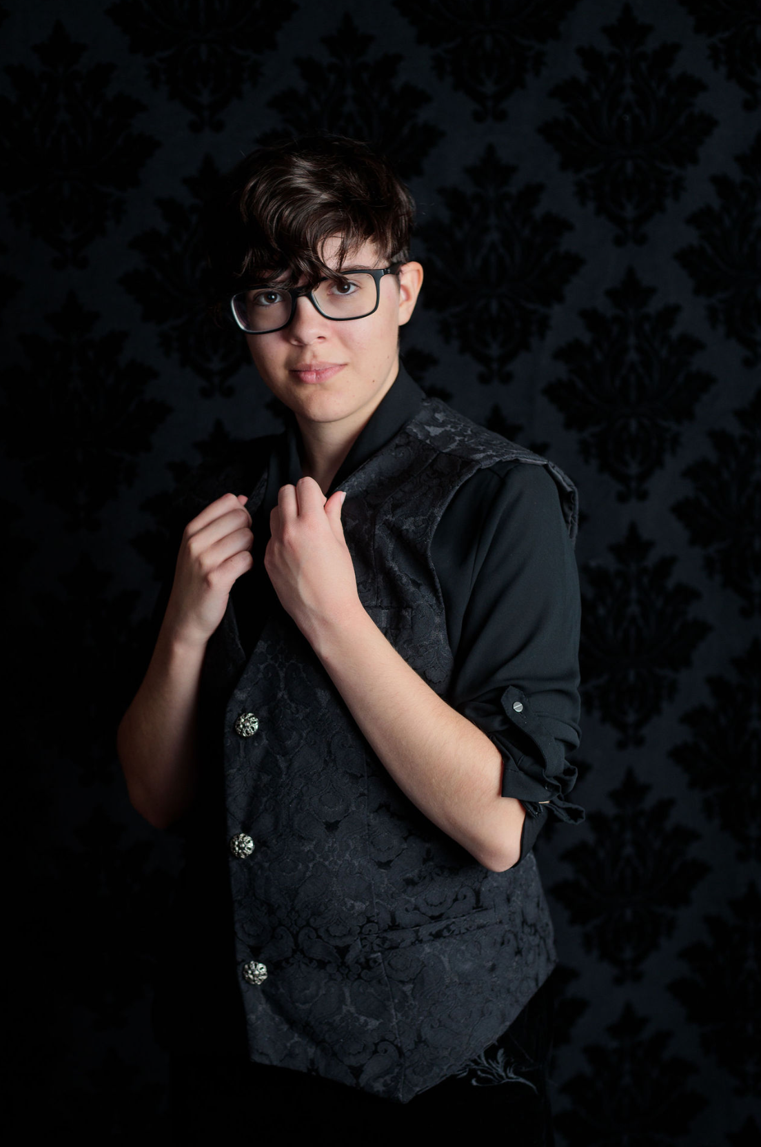 Young person in black brocade vest and glasses posing against dark damask wallpaper background.