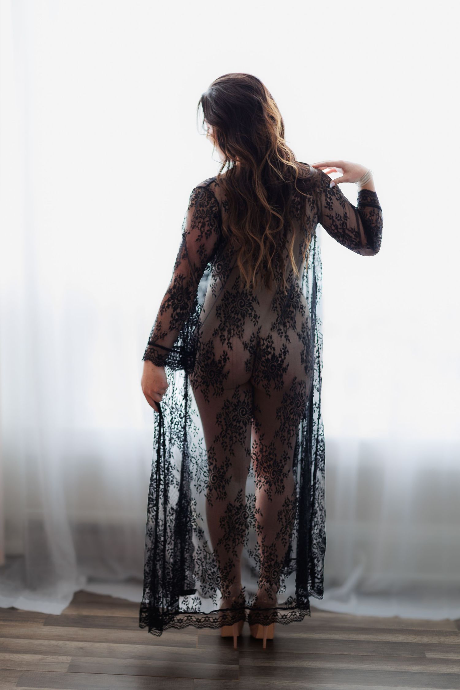 Woman in sheer black lace floral maxi robe standing by bright window, back view, long wavy hair.
