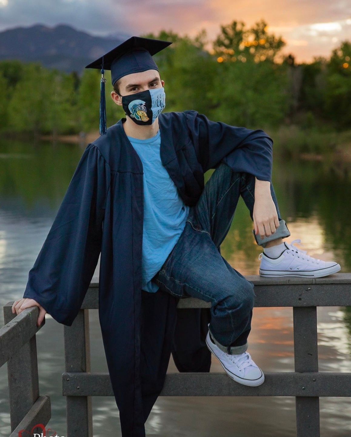 Graduate in cap, gown, and mask poses on wooden dock by lake at sunset.
